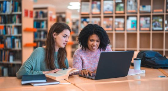 Female Hispanic professor and a Caucasian student sitting in a library studying. Sitting at a desk using laptop as a source of information.